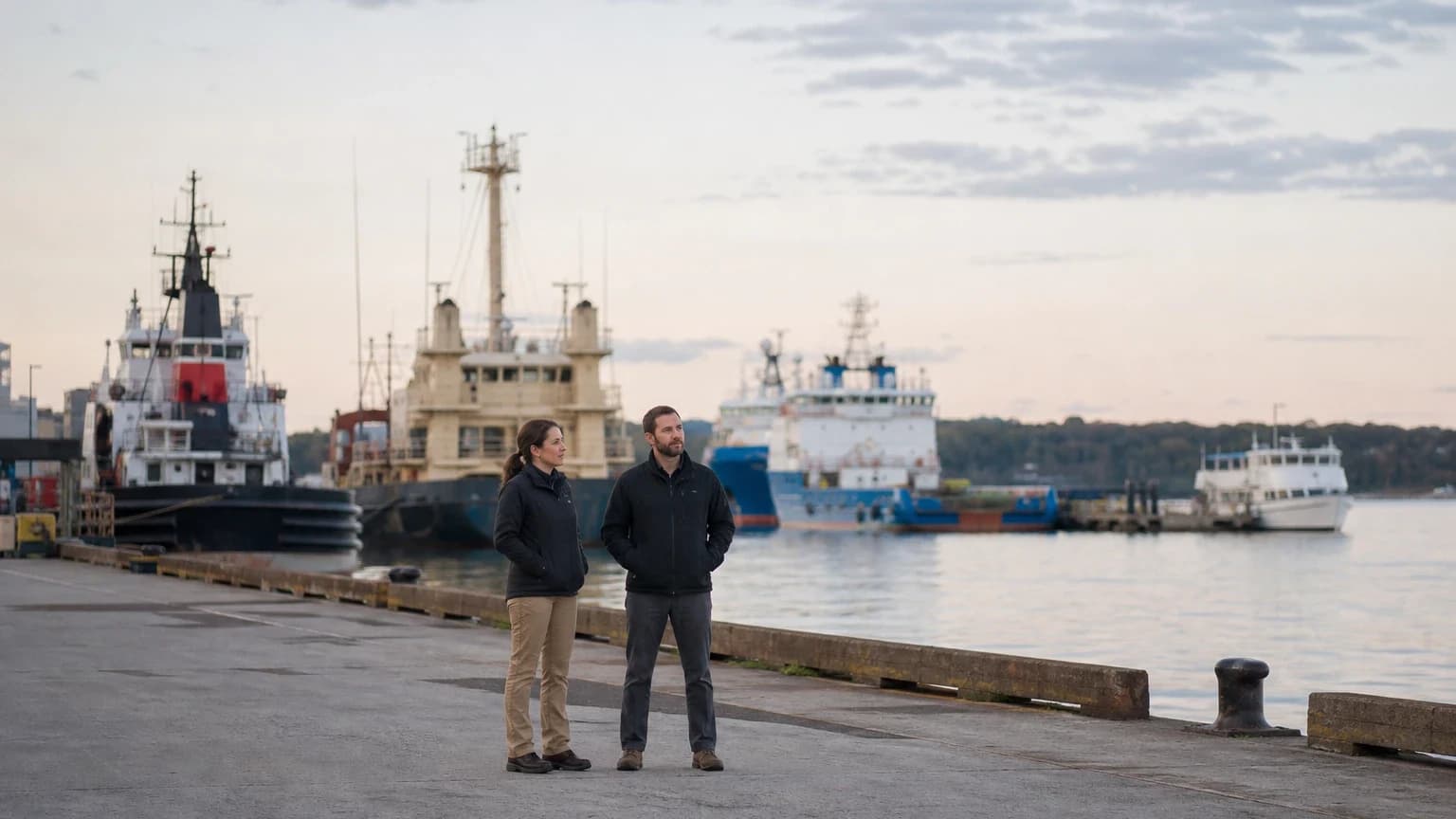 Two fleet compliance professionals stand on a pier with several commercial vessels moored behind them